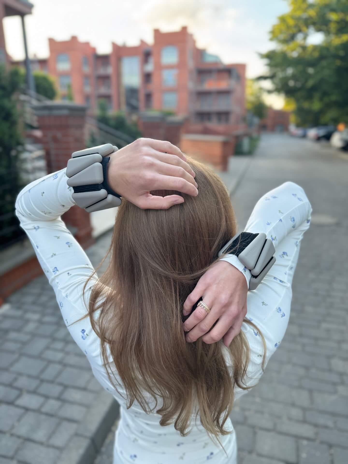 Person with bulky gray robotic wrist braces on both wrists, adjusting hair on a city sidewalk outside.