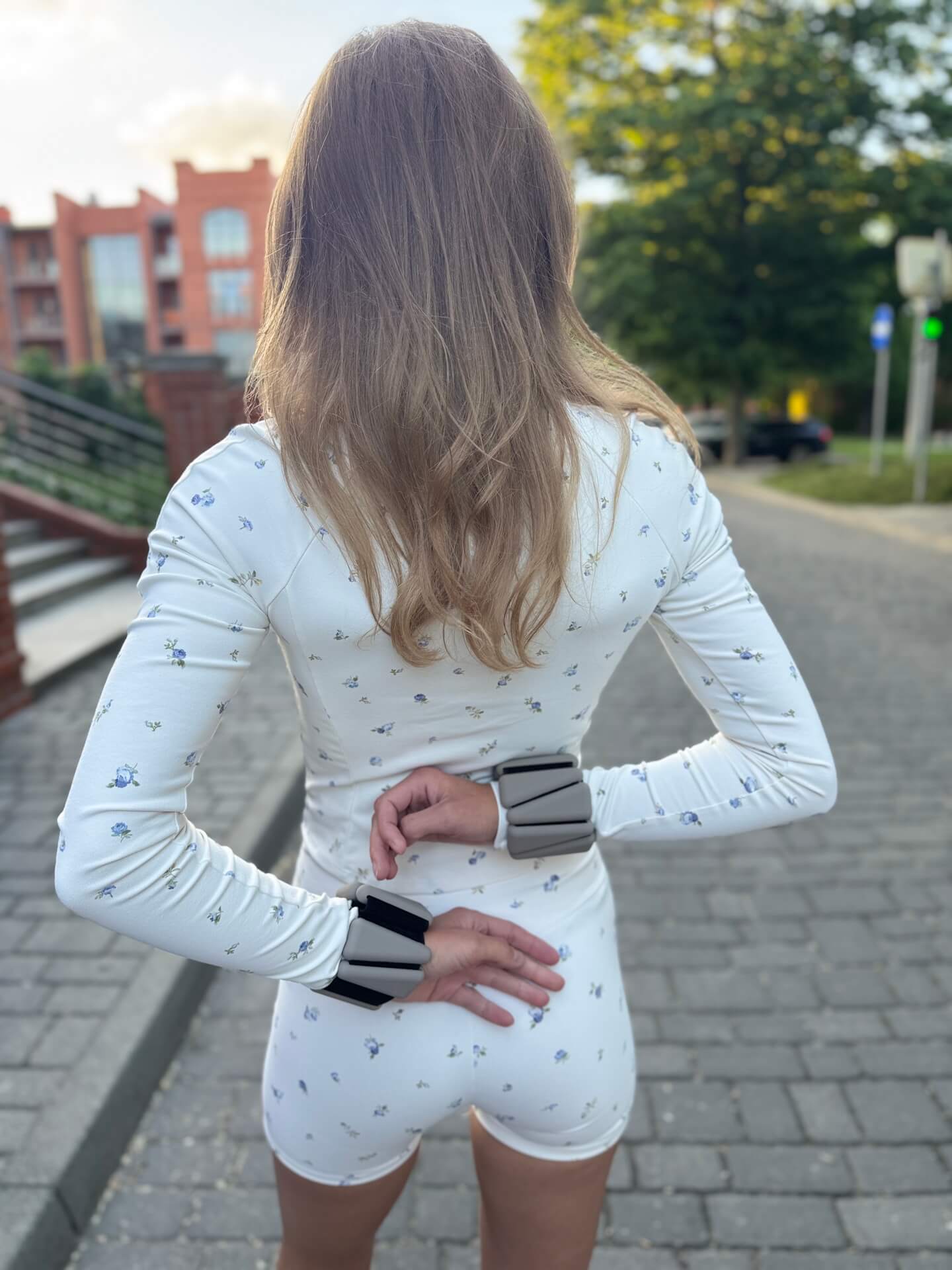 Person seen from behind wearing a white floral outfit, hands on hips, standing on a brick sidewalk outdoors.