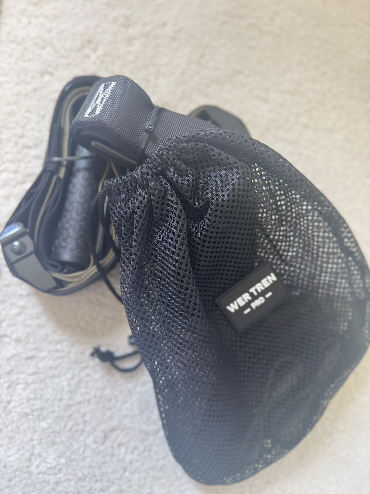 Close-up of a black mesh drawstring bag with a small label reading 'WER TREN' attached to a strap on a light concrete surface